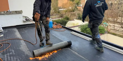 un ouvrier chauffe une membrane derbigum sur une toiture plate avec un chalumeau, un autre ouvrier marche à côté avec un pull siglé Dav toiture